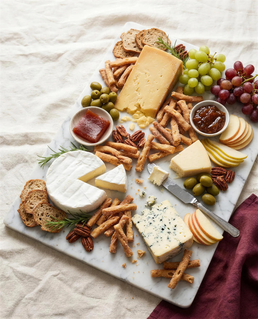 A cheese board on marble with golden Kurma sticks woven among the cheeses, quince paste, olives and grapes, soft daylight