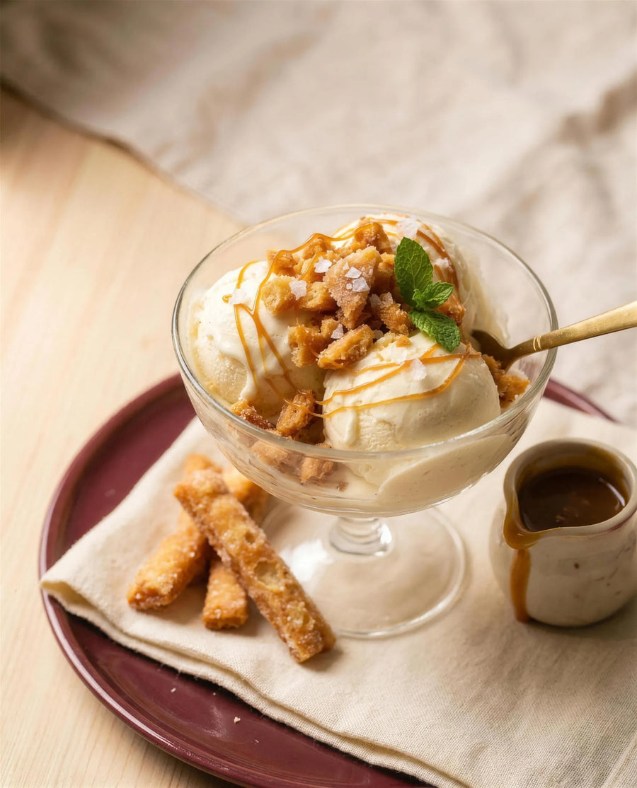 A coupe of vanilla ice cream topped with crushed golden Kurma and a thread of caramel, whole Kurma sticks beside it